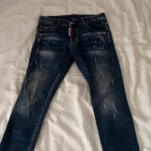 Dsquared2 blå jeans med slitningar - Snygga blå jeans från Dsquared2 med coola slitningar och tvättade detaljer. Klassisk femficksmodell med en tydlig grön Dsquared2-label bak i midjan och röd logga vid gylfen. Jeansen har en smal passform och är tillverkade i jeansmaterial.