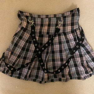 Rutig kortkjol med svarta remmar - Supercool rutig kortkjol i svart, grått och ljusrosa med svarta bältesremmar med silverringar och nitar. Kjolen har plisserad form och ger en edgy vibe, perfekt för dig som gillar alternativ stil. Köpt i USA på Hot Topic och använd en gång. 