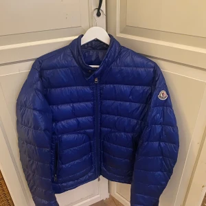 Blå pufferjacka från Moncler - Säljer en blå pufferjacka från Moncler med quiltad design och dragkedja framtill. Jackan har lång ärm och klassisk Moncler-logga på ena ärmen. Perfekt för kyligare dagar. 