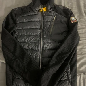 Parajumper cardigan  - Säljer en svart hybridjacka från Parajumpers med quiltade partier och stretchiga ärmar. Jackan har dragkedja framtill, bröstficka med dragkedja och klassisk PJS-logga på ärmen. Perfekt för lager-på-lager under kyliga dagar.