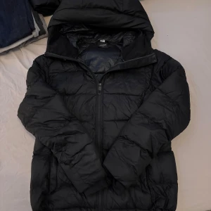 Svart pufferjacka från Everest - Säljer en svart pufferjacka från Everest med huva och dragkedja framtill. Jackan är vadderad och har en klassisk pufferlook som håller dig varm under kyliga dagar. Perfekt för dig som vill ha en enkel och stilren vinterjacka.