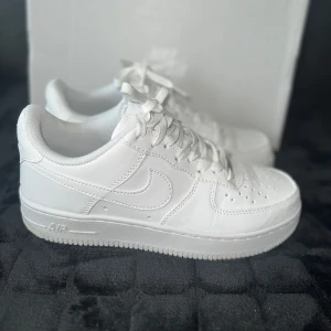 Nike Air Force 1 - Klassiska Nike Air Force 1 i helvitt skinn med vit sula och snörning. Ikonisk design med perforerad tå och broderad Nike-logga på hälen. Perfekta för dig som vill ha en clean och tidlös look till vardags. Levereras med originalkartong.