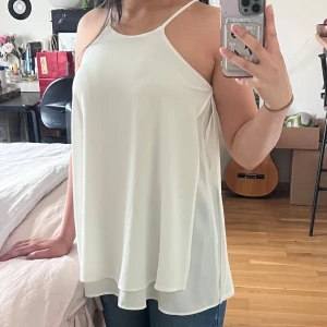 Vit linnetopp från River Island - Säljer en vit, flowy linnetopp från River Island med tunna axelband och lös passform. Toppen har en lätt textur i tyget och är perfekt för varma dagar. Enkel och stilren design som är lätt att matcha med jeans eller kjol.