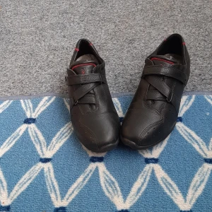 Svarta ECCO sneakers i skinn - Snygga svarta sneakers från ECCO i mjukt skinn med diskreta röda detaljer och kardborreband över vristen. Skorna har rund tå och platt sula, perfekt för dig som gillar stilren och bekväm design. Passar till det mesta och är lätta att ta på.