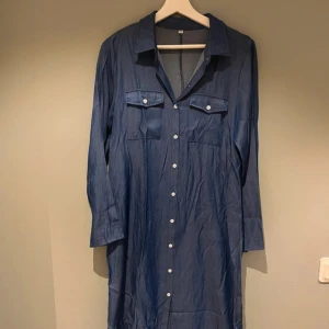 Mörkblå lång skjortklänning med knappar - Snygg mörkblå midiklänning i jeansliknande material. Klänningen har långa ärmar, klassisk krage, vita knappar hela vägen och två bröstfickor. Perfekt för en chill och stilren look. Faller väldigt fint och elegant på. 