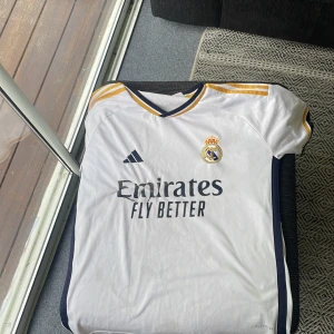Real Madrid hemmatröja Adidas - Snygg vit Real Madrid hemmatröja från Adidas med gula och mörkblå detaljer.  Den är ifrån Säsongen 2023/ 2024.Tröjan har korta ärmar, klubbmärke på bröstet och 'Emirates Fly Better' tryckt på framsidan. Materialet är lätt och andas, perfekt för fotboll eller träning.
