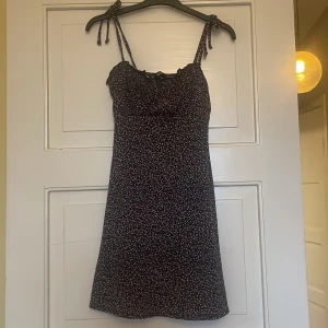 Svart kortklänning med blommigt mönster från H&M Divided - Söt svart kortklänning från H&M Divided med små rosa blommor och smala axelband som knyts. Klänningen har en figurnära passform och är perfekt för varma dagar. Lätt och bekväm att bära.