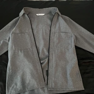 Grå kavajjacka från Zara - Stilren grå kavajjacka från Zara med två stora bröstfickor och dragkedja framtill. Jackan har en klassisk krage och är tillverkad i ett mjukt material som ger en dressad men avslappnad look.