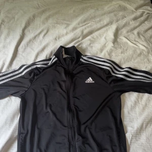 Svart kofta från Adidas - Klassisk svart träningskofta från Adidas med vita ränder längs ärmarna och dragkedja framtill. Koftan har hög krage och Adidas-logga på bröstet. Perfekt för sportiga outfits eller chill vardagsstil.