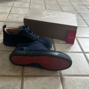  Christian Louboutin skor - Säljer ett par mörkblå sneakers från Christian Louboutin med klassisk röd sula och diskret logga på hälen. Skorna har snörning och är tillverkade med mocka. Kommer med originalkartong och dustbag. Liten skada på mockan på ena tån. Priset är inte hugget i sten och kan ändras