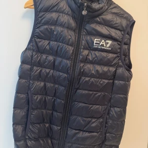 Svart dunväst från Emporio Armani EA7 - Snygg mörkblå dunväst från Emporio Armani EA7 med quiltad design och dragkedja framtill. Västen har en hög krage och logga på bröstet. Perfekt för lager-på-lager och stilren look.