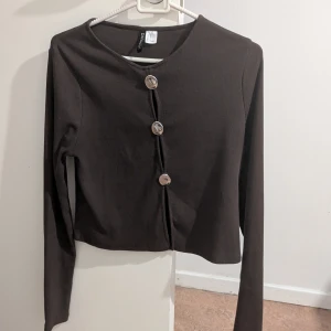 Brun ribbad kofta från H&M - Säljer en brun ribbad kofta från H&M med stora silvriga knappar framtill. Modellen är croppad och har långa ärmar. Perfekt att styla över ett linne eller t-shirt.