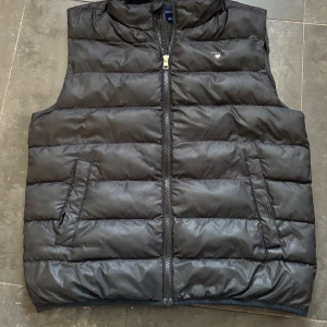 Svart väst från GANT - Säljer en svart dunväst från GANT med dragkedja framtill och två snedställda fickor. Västen har en klassisk, vadderad design och hög krage. Perfekt för lager-på-lager och enkel att matcha med olika outfits. Storleken är 176.