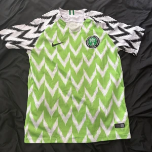 Nigeria fotbollströja VM 2018 - Säljer en officiell Nigeria fotbollströja från VM 2018. Tröjan är kortärmad med ett grönt och vitt zigzag-mönster, svart Nike-logga och Nigeria Football Federation-märke på bröstet. Tillverkad i lätt och ventilerande material. Cond 9/10.
