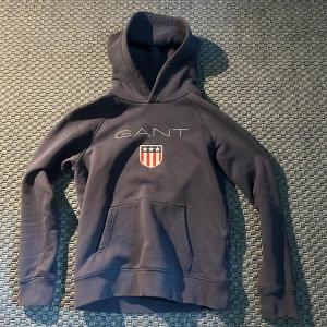 Mörkblå hoodie från GANT - Säljer en mörkblå hoodie från GANT med broderad logga och sköld på bröstet. Tröja köptes för 600 kr och är i storlek 152.