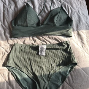 Ribbad grön bikini från H&M - Underdelen Endast provad med trosor på för stor för mig. Överdel 44( hm storlek, dvs liten)  vadderad.  underdel stk 40.  ribbad bikini från H&M i en dämpad grön nyans. Överdelen har trekantskupor med smala axelband och bred resår under bysten. Underdelen är high waist och ger en trendig siluett. Perfekt för strandhäng eller poolparty i sommar!