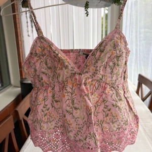 Rosa blommig linnetopp från Bikbok - Supersöt rosa linnetopp från Bikbok med blommigt mönster i grönt, vitt och gult. Toppen har smala axelband, v-ringning och broderade volangdetaljer längst ner. Perfekt för varma dagar och ger en romantisk vibe.