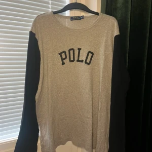 Grå långärmad tröja från Polo Ralph Lauren - Snygg långärmad tröja från Polo Ralph Lauren i grått med svarta ärmar och svart POLO-tryck på bröstet. Tröjan har en avslappnad passform och är gjord i mjuk bomull, perfekt för en chill och stilren look.