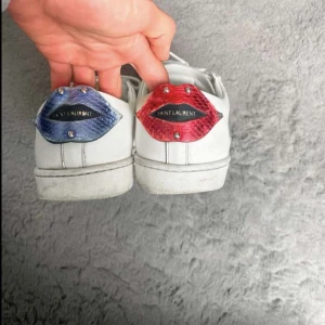 Vita Saint Laurent sneakers - Unika vita sneakers från Saint Laurent med snörning och coola läppar i blått och rött på hälen. Skorna har rund tå och platt sula, tillverkade i skinn. Perfekta för dig som vill sticka ut med en exklusiv touch.