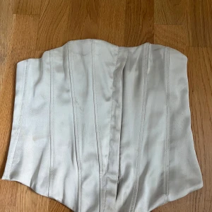 Vit korsett från Zara - Säljer en stilren vit korsett från Zara. Modellen är axelbandslös och har markerade sömmar för en snygg passform. Perfekt att styla till olika outfits. Stängs med hyskor på sidan.