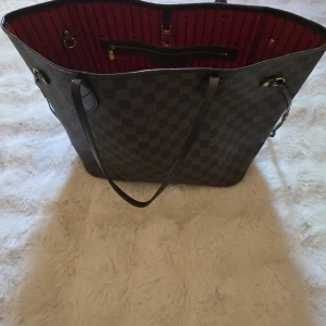 Neverfull från Louis Vuitton - Säljer en klassisk Neverfull från Louis Vuitton i deras ikoniska rutiga mönster. Väskan har rödrandigt foder, dragkedjeficka på insidan och långa axelband. Perfekt för dig som vill ha en rymlig och stilren accessoar. Se bilder för att se skick!