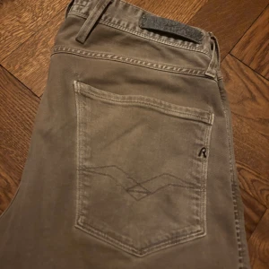Replay Anbass grå jeans slim fit - Snygga grå Replay Anbass jeans i slim fit med klassisk femficksdesign och diskret logga på fickan. Byxorna har normal midja och raka ben, tillverkade i mjuk bomull med lite stretch för extra komfort. Perfekta för en clean och stilren look.