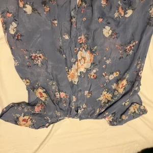 Blommig blå blus från KappAhl - Supersöt blå blus från KappAhl med blommigt mönster i pastellfärger. Lätt och flowy känsla, knappar framtill och mjukt tyg som passar perfekt för varmare dagar. Perfekt för dig som gillar romantisk och feminin stil.