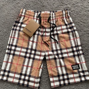 Rutig shorts från Burberry - Snygga shorts från Burberry med klassiskt rutigt mönster i beige, svart, vitt och rött. De har elastisk midja med svart snörning och en Burberry London England-patch framtill. Perfekta för sommaren och riktigt trendiga.