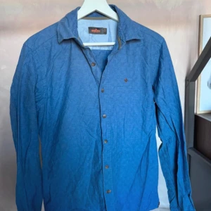 Blå skjorta från Morris - Snygg blå skjorta från Morris med klassisk krage och knappar framtill. Skjortan har lång ärm och en liten broderad detalj på bröstet. Perfekt för dig som gillar stilrena plagg med diskreta detaljer.