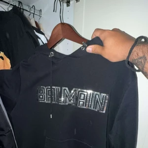 Svart Balmain hoodie med silverdetaljer - Säljer en svart hoodie från Balmain med stort glittrigt silvertryck på bröstet. Tröjan har huva med snörning och en klassisk känguruficka framtill. Perfekt för dig som vill sticka ut med en exklusiv streetstyle-look.