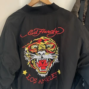 Svart bomberjacka från Ed Hardy - Snygg svart bomberjacka från Ed Hardy med ett stort broderat tigerhuvud och texten 'Los Angeles' på ryggen. Avslappnat stil!! Aldrig använd och säljer då den tyvärr inte passar mig. 