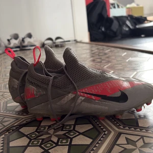Grå och röda fotbollsskor från Nike - Snygga fotbollsskor från Nike i grått med röda detaljer och en svart swoosh. Skorna har en strömlinjeformad design med snörning och dragflikar för enkel påtagning. Perfekta för fotbollsplanen med sin greppvänliga sula.