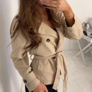 trenchcoat  - Snygg beige trenchcoat med knytbälte och knappar. Perfekt för vår och höst. Jackan har långa ärmar och en klassisk design som passar till många olika stilar.