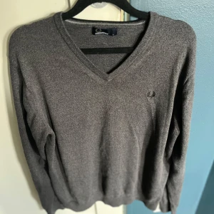 Grå tröja från Fred Perry - Säljer en stilren grå tröja från Fred Perry med v-ringning. Tröjan har långa ärmar och en diskret logga på bröstet. Perfekt för en klassisk och avslappnad look. Sitter som en M