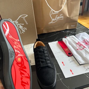 Svarta sneakers från Christian Louboutin - Snygga svarta sneakers i mocka från Christian Louboutin med den ikoniska röda sulan. Skorna har snörning och en rund tå, vilket ger en stilren look. Perfekta för dig som vill ha en touch av lyx i vardagen.