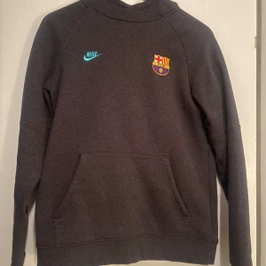 Grå hoodie från Nike med FC Barcelona-logga - Säljer en grå hoodie från Nike med FC Barcelona-logga på bröstet. Tröjan har en klassisk känguruficka och Nike-logga i turkos. Perfekt för fotbollsfans som vill hålla sig varma och stiliga. Storleken är XL men känns som S.
