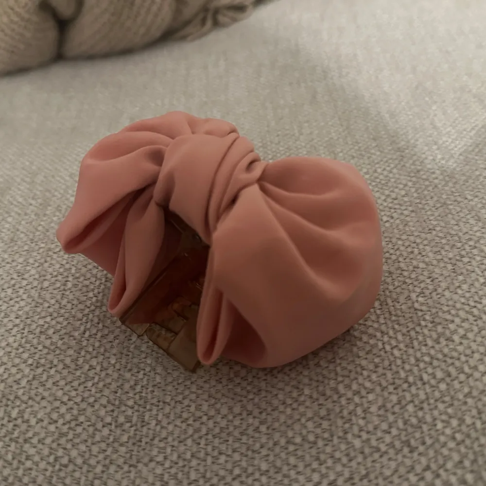 Söt rosa hårklämma med en stor rosett. Perfekt för att ge en extra touch till din frisyr. Klämman är gjord av plast och har en mjuk, tygklädd yta.. Asusteet.