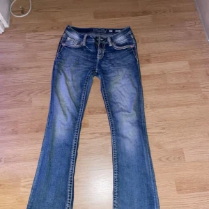 Miss Me Blå bootcut jeans med broderade fickor - Snygga blå bootcut jeans med coola broderade detaljer på bakfickorna. Jeansen har en klassisk femficksdesign och en lätt slitning för en trendig look. Perfekta för en avslappnad stil. Lagning vid skrev som it syns på