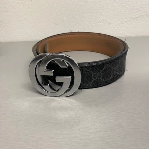 Svart bälte  - Snyggt svart bälte med ett klassiskt GG-mönster och en stor silverfärgad logotypspänne. Tillverkat i Italien med högkvalitativt material. Perfekt för att ge en lyxig touch till din outfit.