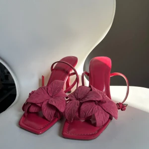 Rosa sandaler med blommig detalj - Snygga rosa sandaler med en stor blommig detalj på ovansidan. De har en elegant klack och smala remmar som ger en feminin touch. Perfekta för att lyfta vilken outfit som helst.
