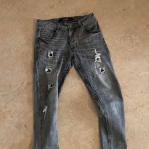 Grå jeans från Alessandro Zavetti - Säljer ett par grå jeans från Alessandro Zavetti med slitna detaljer och en cool, distressed look. Byxorna har en klassisk femficksdesign och knappgylf. Perfekta för en avslappnad stil.