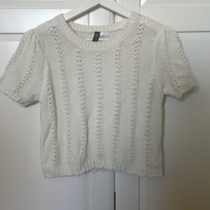 Vit stickad kortärmad tröja från H&M - Säljer en söt vit stickad tröja från H&M med korta ärmar och ett fint hålmönster. Perfekt för vår och sommar. Tröjan har en rund halsringning och ribbade kanter.