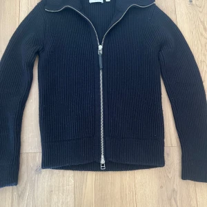 Svart ribbad kofta med dragkedja - Snygg svart ribbad kofta från Tiger of sweden med dragkedja framtill. Perfekt för kyligare dagar med sin stilrena design och långa ärmar. Passar bra till både jeans och chinos. PRIS KAN DISKUTERAS 