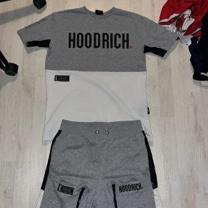 Hoodrich T-shirt och shorts - Snygga grå shorts från Hoodrich med svart logga på benet. Shortsen har dragkedjor på fickorna och en elastisk midja för extra komfort. Perfekta för en avslappnad stil.