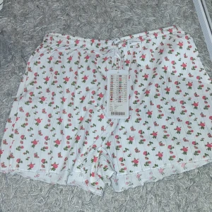Vita shorts med blommigt mönster - Söta vita shorts med ett blommigt mönster i rosa och grönt. De har en bekväm passform och elastisk midja med knytband. Perfekta för en avslappnad sommardag. Har fortfarande prislappen kvar (kostade från början 95)