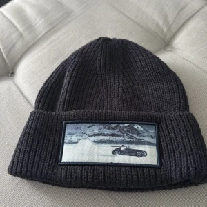 Svart ribbad mössa med bilmotiv - Snygg svart ribbad mössa med ett unikt bilmotiv på framsidan. Perfekt för att hålla värmen under kyliga dagar. Stilren och enkel design som passar till det mesta.