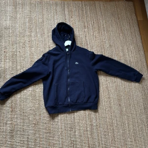 Mörkblå hoodie från Lacoste - Säljer en stilren mörkblå hoodie från Lacoste med dragkedja och klassisk krokodil-logga på bröstet. Tröjan är i storlek XL men skulle säga att den är en L. Är 196 cm och den är kort i midjan. Har Max använt den 5-6 gånger.