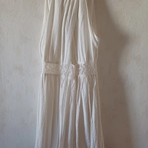 Vit ärmlös klänning med spetsdetaljer - Säljer en elegant vit klänning med vacker spetsdetalj i midjan. Klänningen är ärmlös och har en djup v-ringning med knytband i nacken. Perfekt för sommarens festligheter!