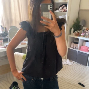 Svart ärmlös topp med spetsdetaljer - Säljer en snygg svart ärmlös topp med vackra spetsdetaljer på axlarna. Perfekt för en stilren look. Toppen har en rund halsringning och en luftig passform. Passar bra till både jeans och kjol. Hör av dig vid intresse!