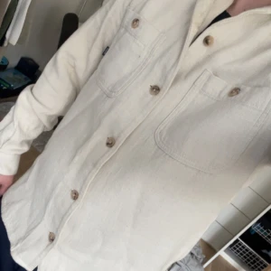 Beige skjorta från Dobber - Säljer en stilren beige skjorta från Dobber i storlek small. Skjortan har långa ärmar och två bröstfickor med knappar. Perfekt för en avslappnad look.